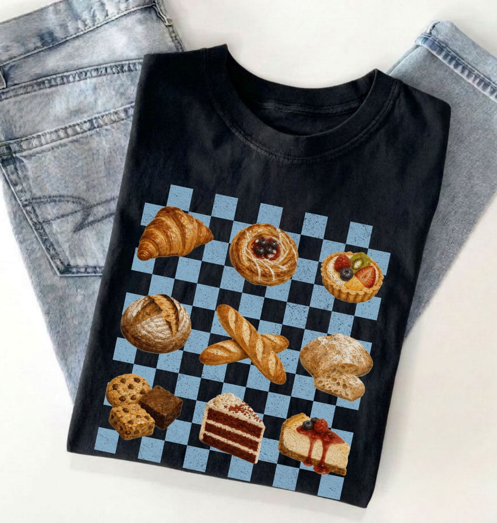 A black short-sleeve T-shirt laid flat on a white surface, styled with light blue jeans at the top. The shirt features a large graphic print of assorted pastries and cakes arranged on a blue-and-white checkered square.