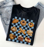 A black short-sleeve T-shirt laid flat on a white surface, styled with light blue jeans at the top. The shirt features a large graphic print of assorted pastries and cakes arranged on a blue-and-white checkered square.