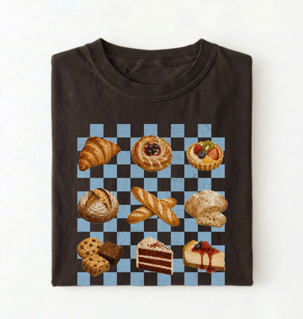 A folded dark brown t-shirt on a white background, featuring a graphic print of various baked pastries and cakes arranged on a black and light blue checkered grid.