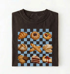 A folded dark brown t-shirt on a white background, featuring a graphic print of various baked pastries and cakes arranged on a black and light blue checkered grid.