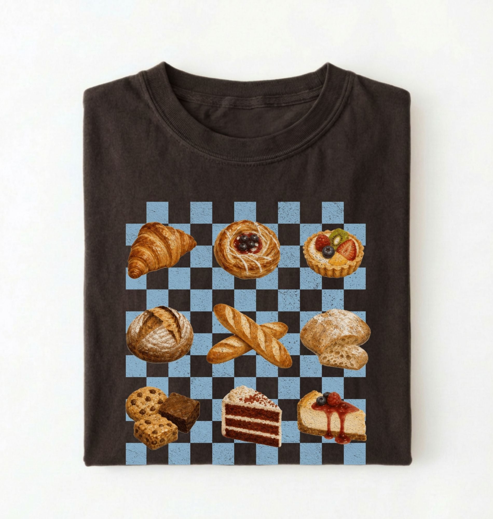A folded dark brown t-shirt on a white background, featuring a graphic print of various baked pastries and cakes arranged on a black and light blue checkered grid.
