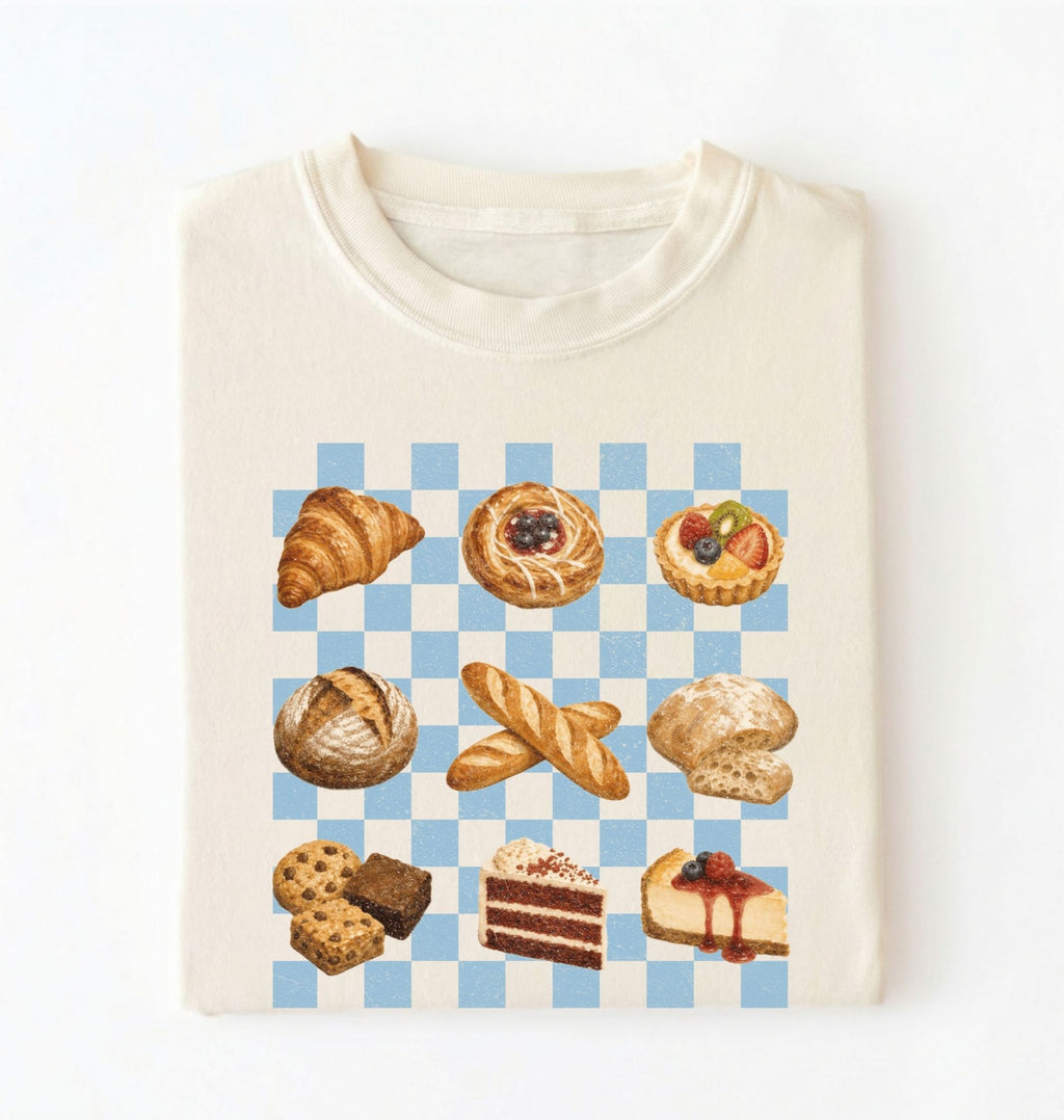 An off-white crewneck t-shirt featuring a graphic print of various baked pastries and cakes arranged on a light blue and white checkered board. The pastries include a croissant, a filled pastry, a tart, a loaf of bread, baguettes, a biscuit, cookies, chocolate cake, and cheesecake.