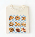 An off-white crewneck t-shirt featuring a graphic print of various baked pastries and cakes arranged on a light blue and white checkered board. The pastries include a croissant, a filled pastry, a tart, a loaf of bread, baguettes, a biscuit, cookies, chocolate cake, and cheesecake.
