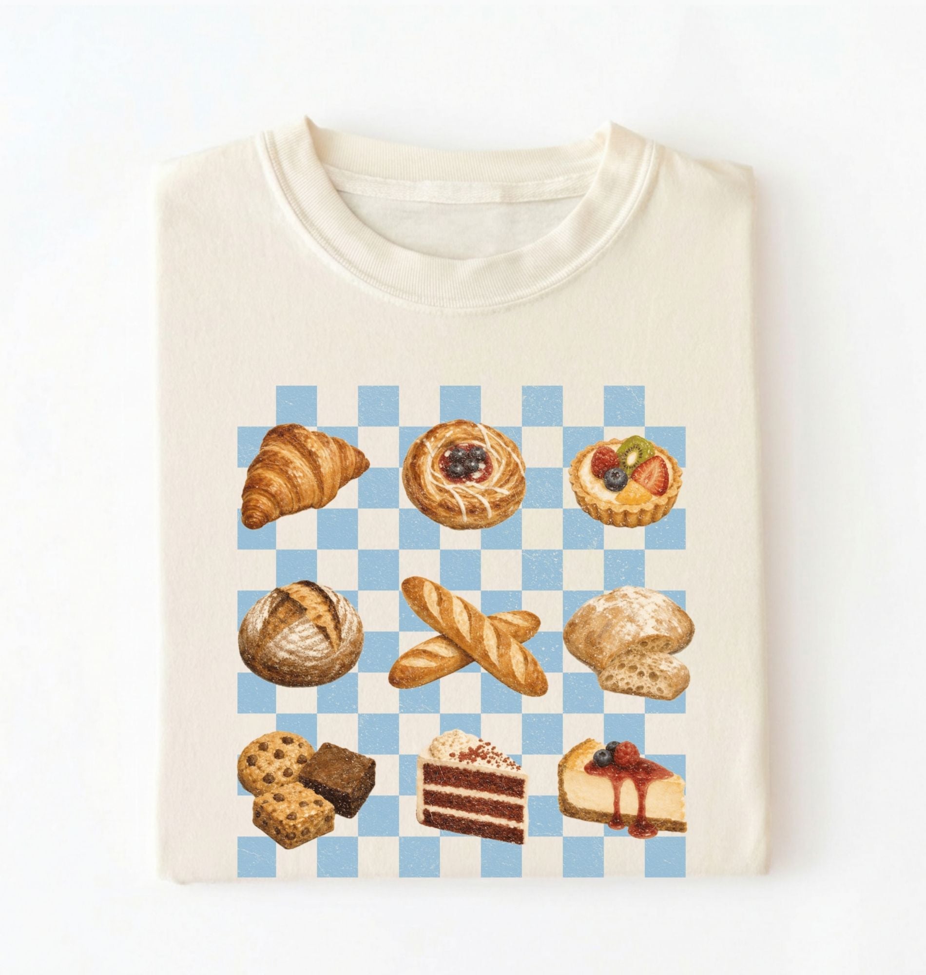 An off-white crewneck t-shirt featuring a graphic print of various baked pastries and cakes arranged on a light blue and white checkered board. The pastries include a croissant, a filled pastry, a tart, a loaf of bread, baguettes, a biscuit, cookies, chocolate cake, and cheesecake.