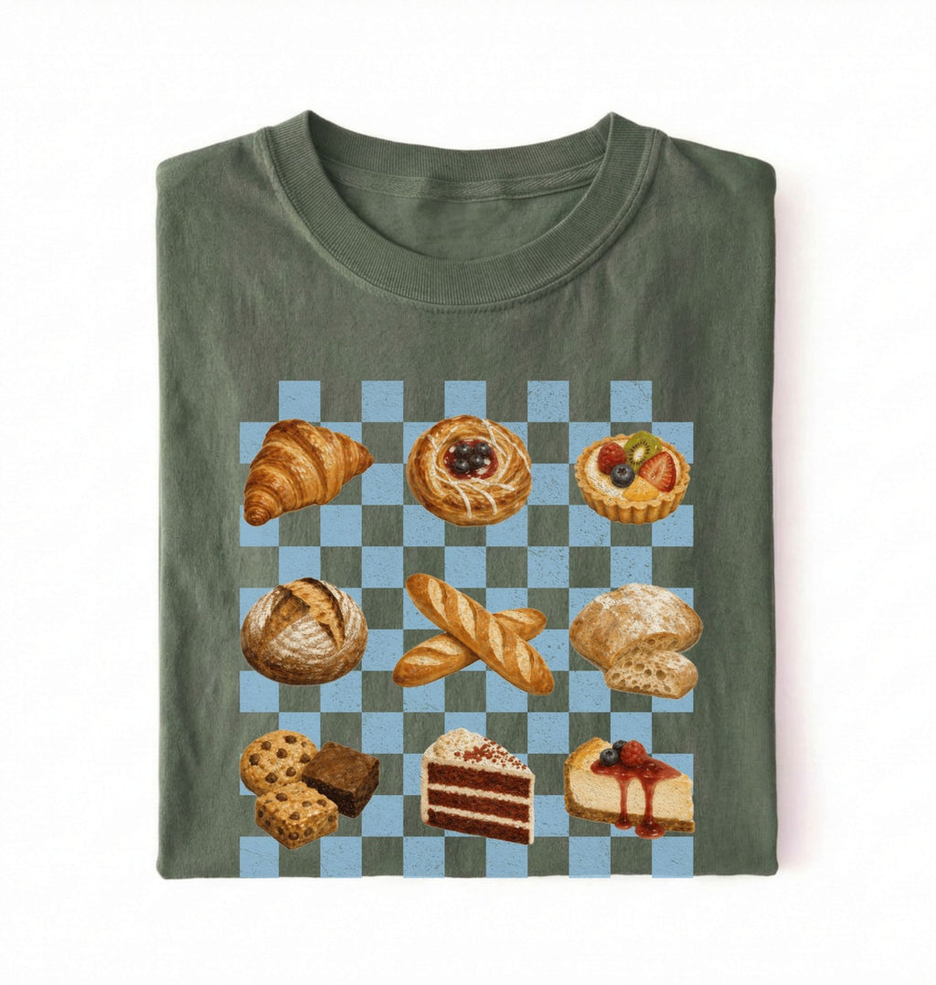 A folded olive green t-shirt with a graphic print of various pastries and cakes arranged on a light blue and gray checkered board, set against a plain white background.