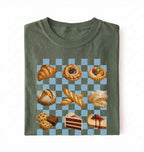 A folded olive green t-shirt with a graphic print of various pastries and cakes arranged on a light blue and gray checkered board, set against a plain white background.