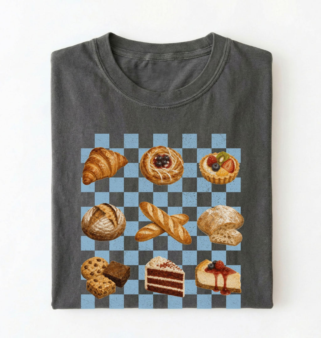 A folded dark gray t-shirt on a white background, featuring a graphic print of various pastries and cakes arranged on a blue and gray checkered board 