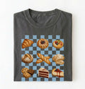 A folded dark gray t-shirt on a white background, featuring a graphic print of various pastries and cakes arranged on a blue and gray checkered board 