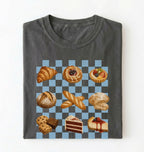 A folded dark gray t-shirt on a white background, featuring a graphic print of various pastries and cakes arranged on a blue and gray checkered board 
