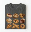 A dark gray crewneck t-shirt featuring a graphic print of various baked goods, including cakes, pastries, cookies, and breads, arranged in a grid on a white background.