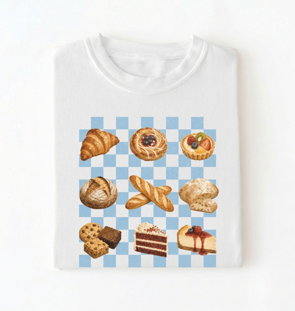 A white crewneck t-shirt with a graphic print of various baked pastries and cakes arranged on a light blue and white checkered background.