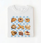 A white crewneck t-shirt with a graphic print of various baked pastries and cakes arranged on a light blue and white checkered background.