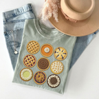 A folded sage green t-shirt featuring embroidered pie designs, styled with light-wash denim jeans, a beige wide-brim hat, and dried pampas grass on a white background.