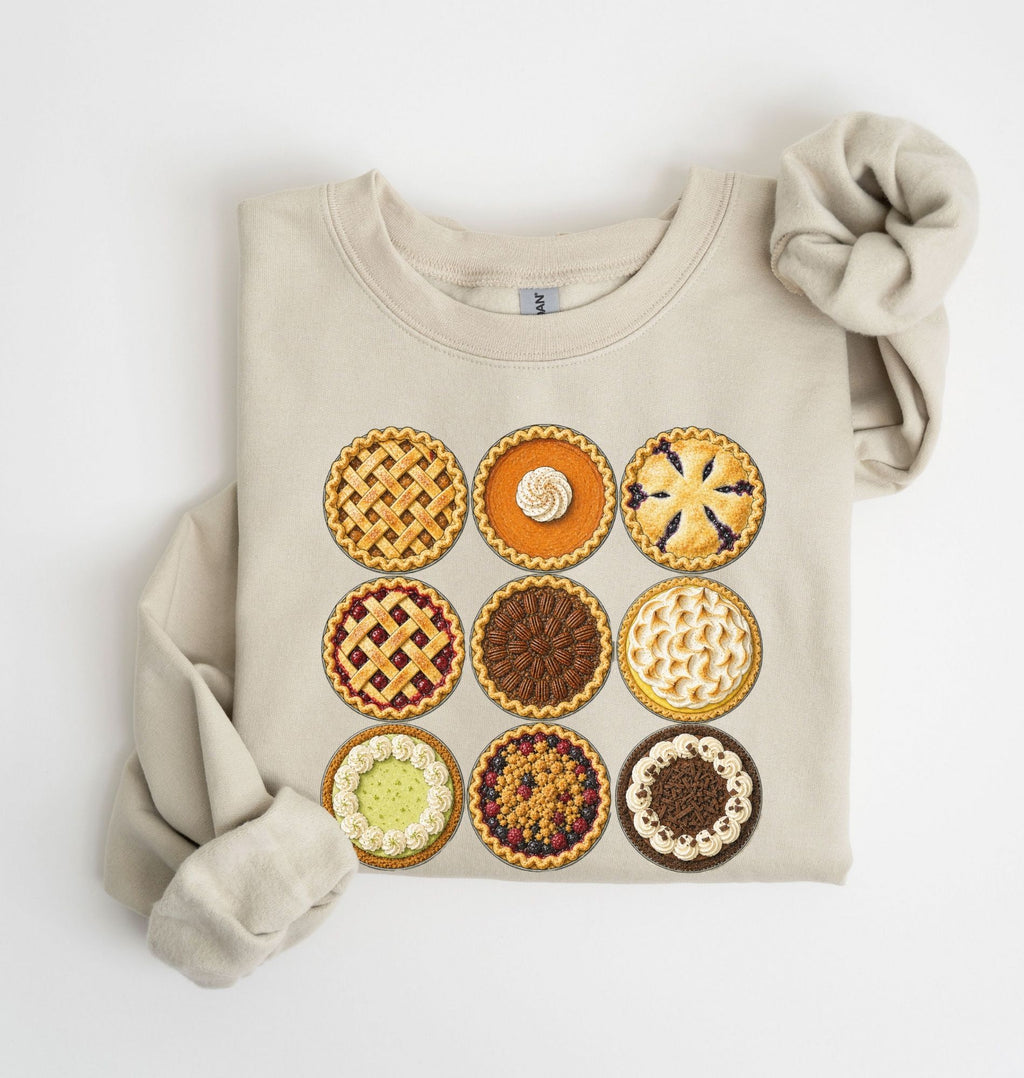 A folded beige sweatshirt with a graphic print of nine different pies arranged in a 3x3 grid, styled with a matching beige knit scrunchie on a white background.