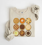 A folded beige sweatshirt with a graphic print of nine different pies arranged in a 3x3 grid, styled with a matching beige knit scrunchie on a white background.