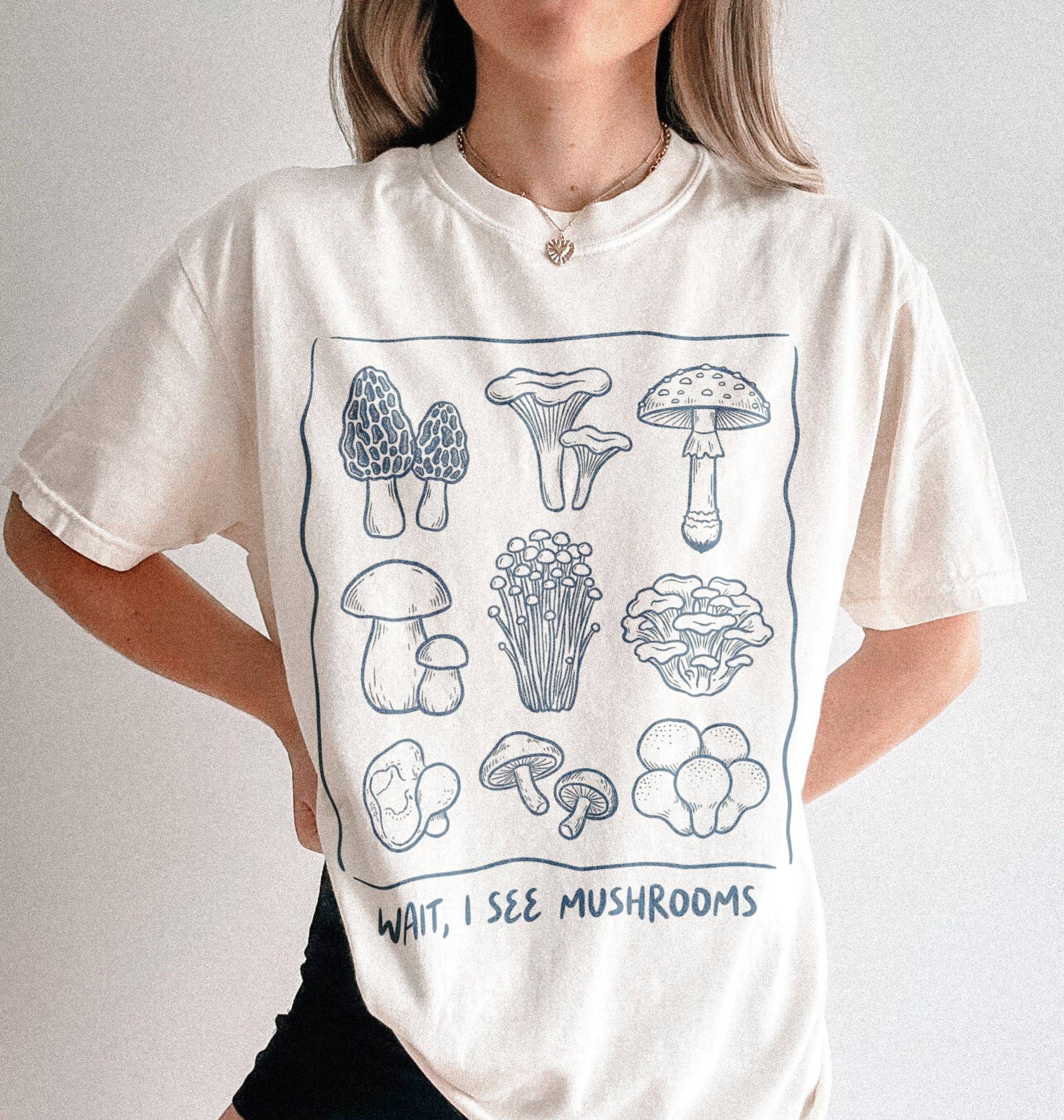 woman wearing a natural cream t shirt with hand drawn mushroom illustrations arranged in a grid and the text wait i see mushrooms underneath