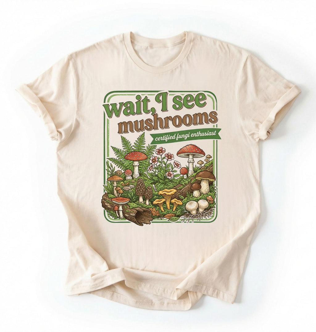 Flat lay of a natural t-shirt featuring illustrated mushrooms and plants with text reading “Wait I See Mushrooms Certified Fungi Enthusiast on a white background