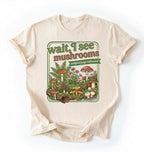 Flat lay of a natural t-shirt featuring illustrated mushrooms and plants with text reading “Wait I See Mushrooms Certified Fungi Enthusiast on a white background