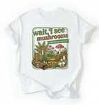 Flat lay of a white t-shirt featuring illustrated mushrooms and plants with text reading “Wait I See Mushrooms Certified Fungi Enthusiast on a white background