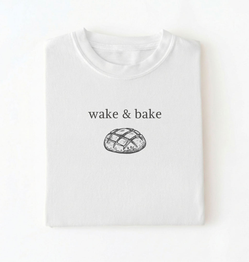 Flat lay of a white Funny wake and bake bread shirt perfect for bakers bread lovers and home baking enthusiasts on a white background
