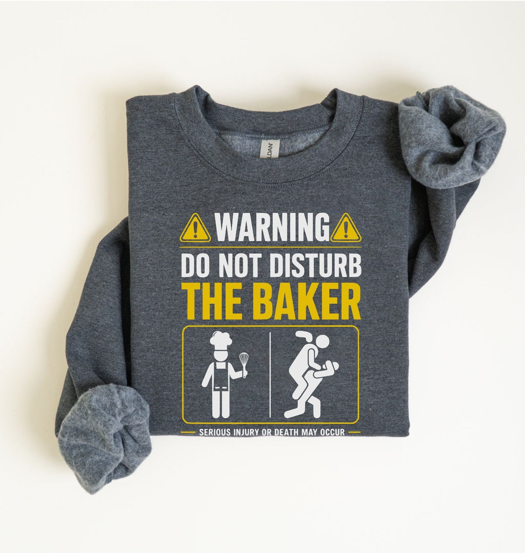 A folded dark heather gray long-sleeve t-shirt with a graphic print that reads 'WARNING DO NOT DISTURB THE BAKER' in white and yellow, accompanied by cartoon illustrations of a baker and a person falling, on a light beige background.