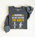 A folded dark heather gray long-sleeve t-shirt with a graphic print that reads 'WARNING DO NOT DISTURB THE BAKER' in white and yellow, accompanied by cartoon illustrations of a baker and a person falling, on a light beige background.