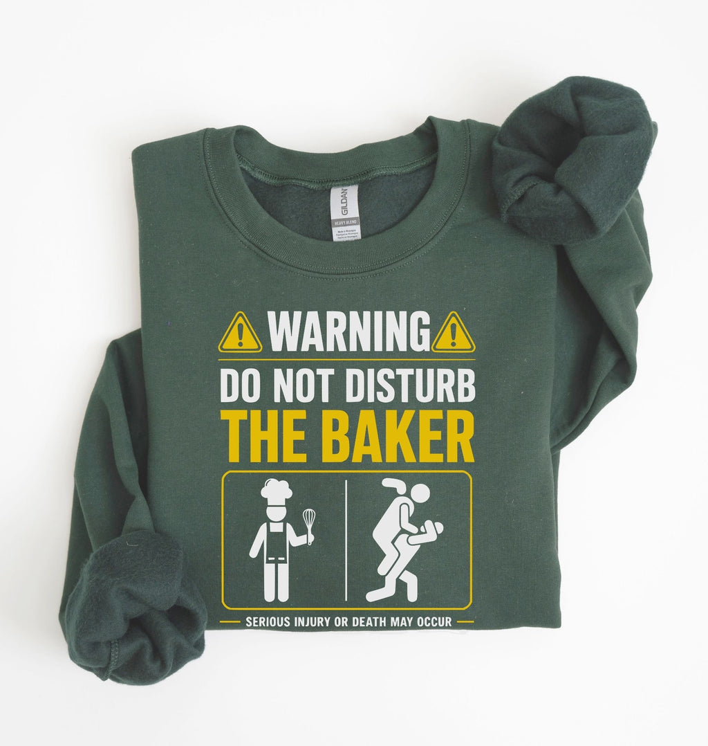 A dark green crewneck sweatshirt with a humorous warning graphic printed on the front. The graphic reads 'WARNING DO NOT DISTURB THE BAKER' in white and yellow text, accompanied by stick-figure illustrations of a baker and a person crouching.