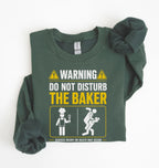 A dark green crewneck sweatshirt with a humorous warning graphic printed on the front. The graphic reads 'WARNING DO NOT DISTURB THE BAKER' in white and yellow text, accompanied by stick-figure illustrations of a baker and a person crouching.