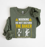 An olive green long-sleeve t-shirt with a humorous warning graphic printed on the front, styled flat on a white background.