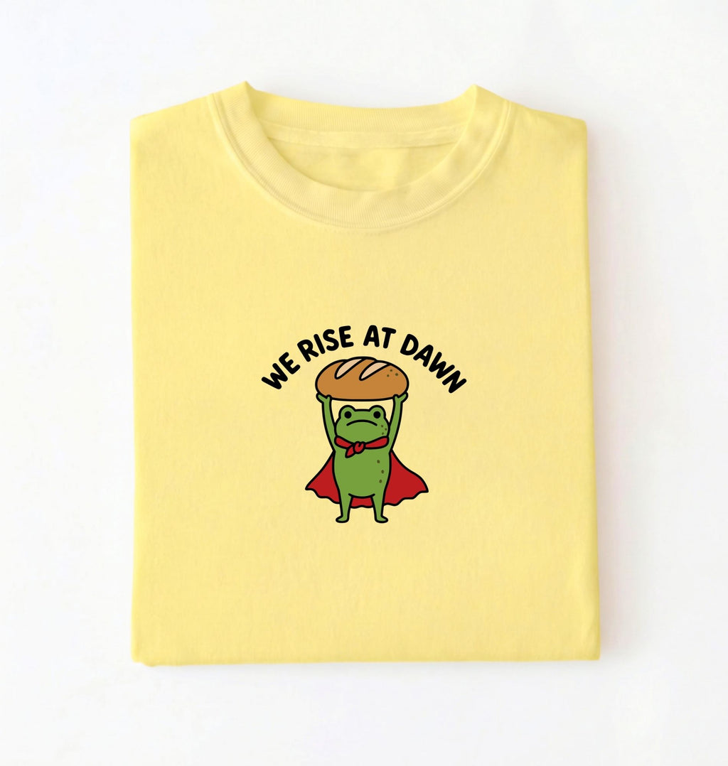 Flat lay of a Cornsilk Funny We Rise At Dawn frog baker shirt featuring bread loaf graphic for sourdough enthusiasts on a white background 