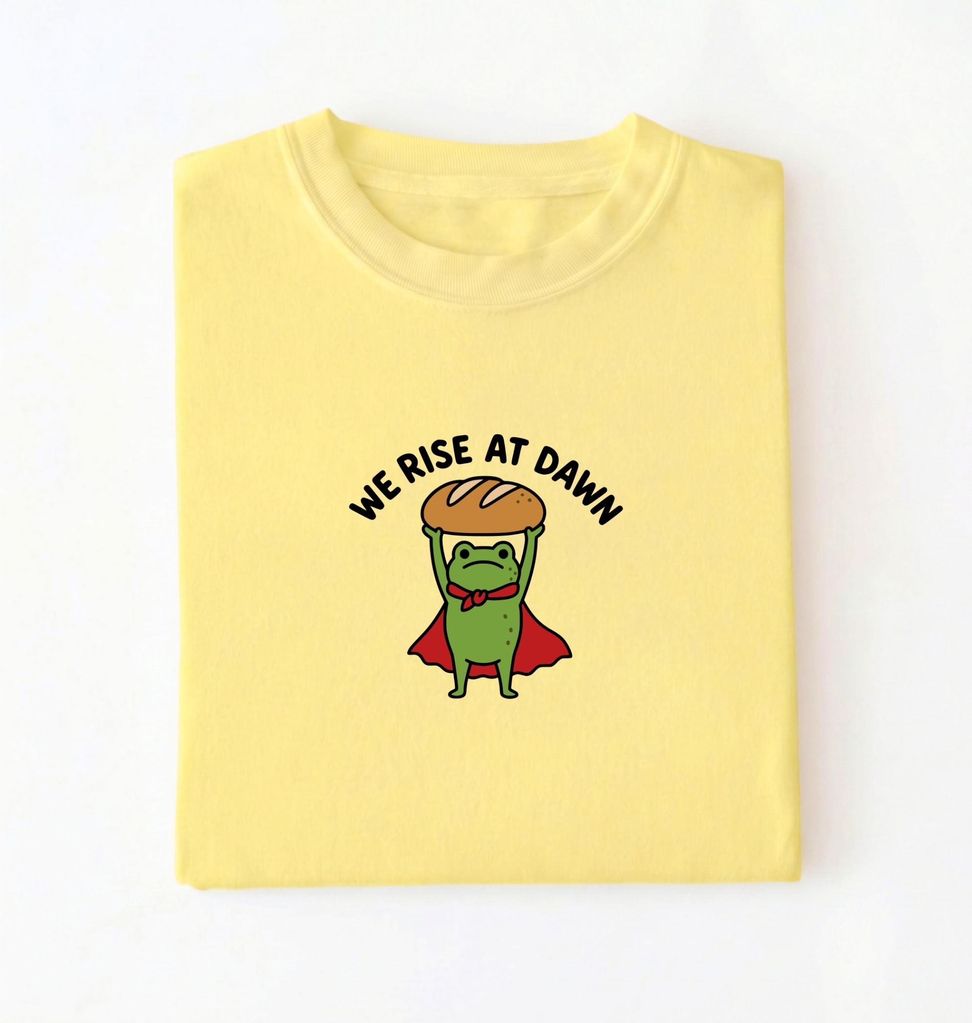 Flat lay of a Cornsilk Funny We Rise At Dawn frog baker shirt featuring bread loaf graphic for sourdough enthusiasts on a white background 