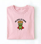Flat lay of a Light Pink Funny We Rise At Dawn frog baker shirt featuring bread loaf graphic for sourdough enthusiasts on a white background 