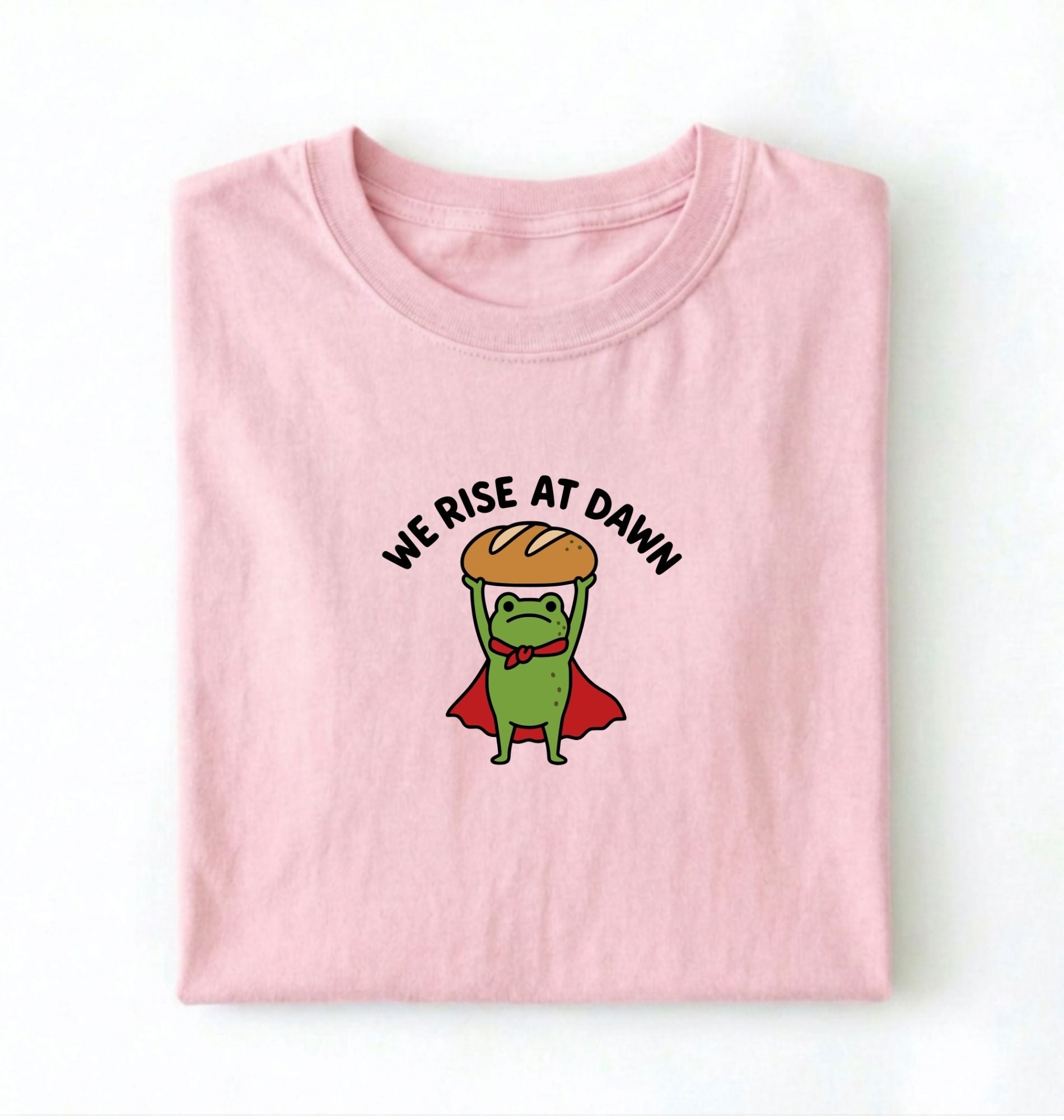 Flat lay of a Light Pink Funny We Rise At Dawn frog baker shirt featuring bread loaf graphic for sourdough enthusiasts on a white background 