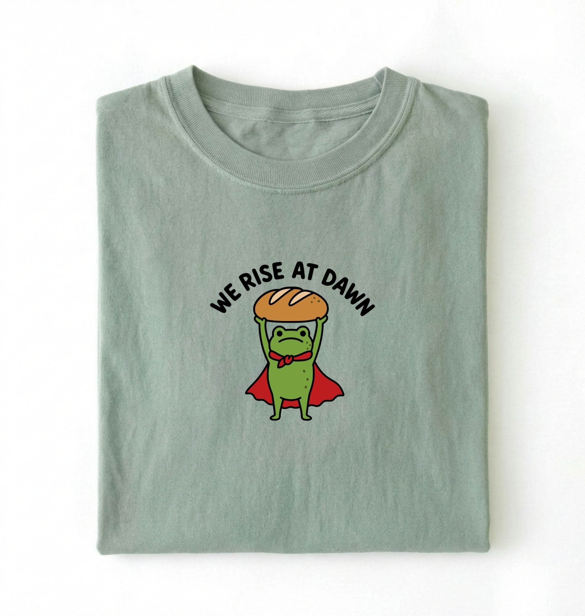 Flat lay of a bay Funny We Rise At Dawn frog baker shirt featuring bread loaf graphic for sourdough enthusiasts on a white background 