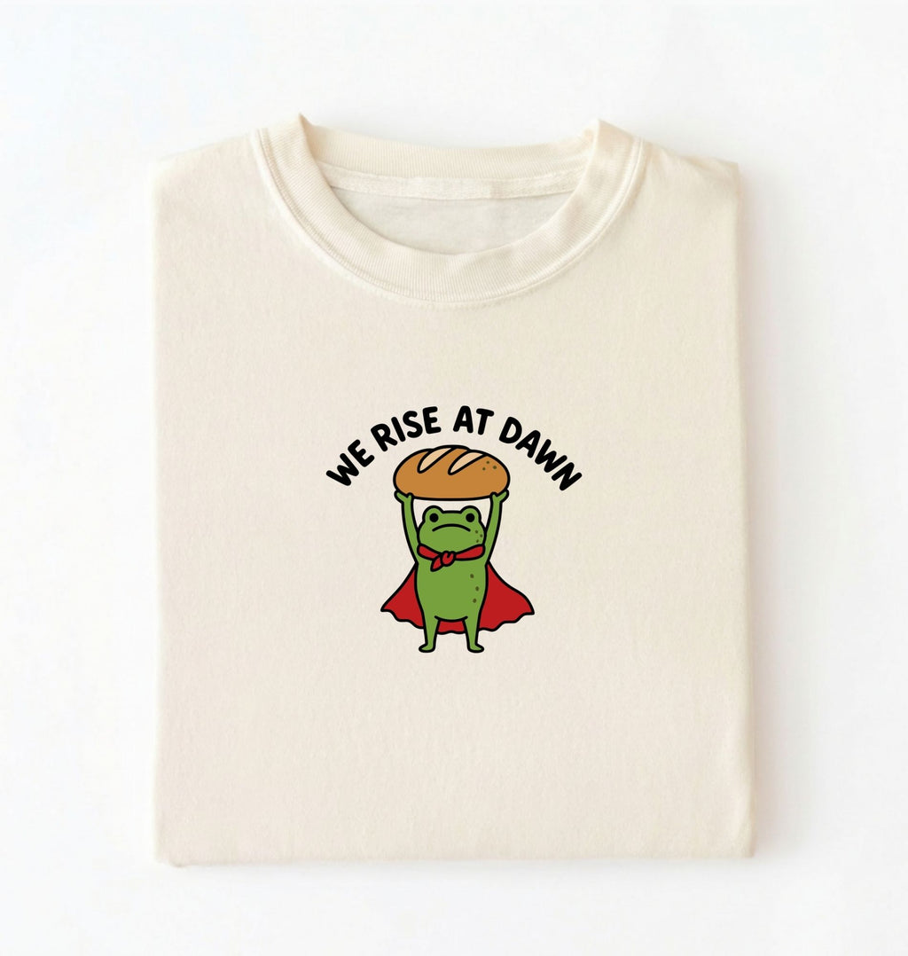 Flat lay of a ivory Funny We Rise At Dawn frog baker shirt featuring bread loaf graphic for sourdough enthusiasts on a white background 