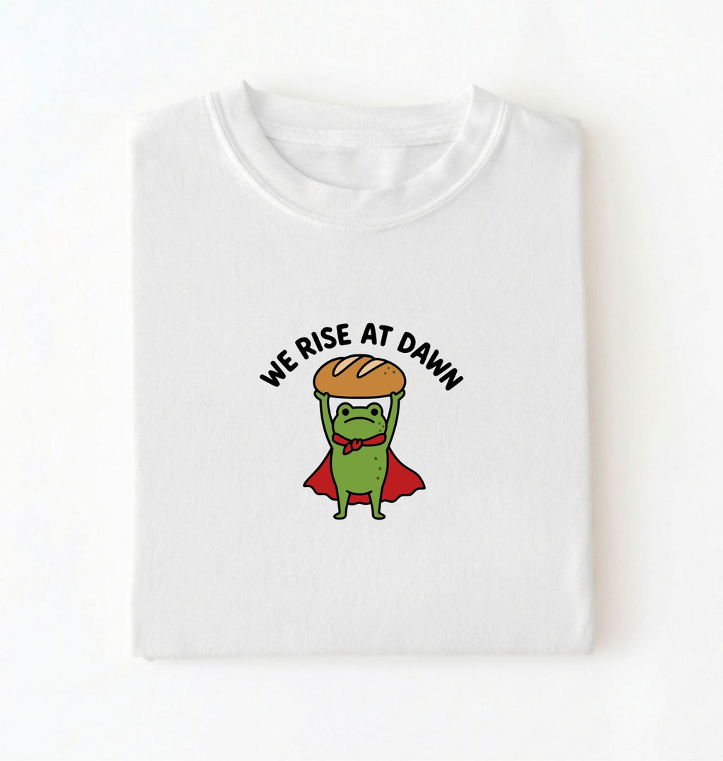 Flat lay of a white Funny We Rise At Dawn frog baker shirt featuring bread loaf graphic for sourdough enthusiasts on a white background 
