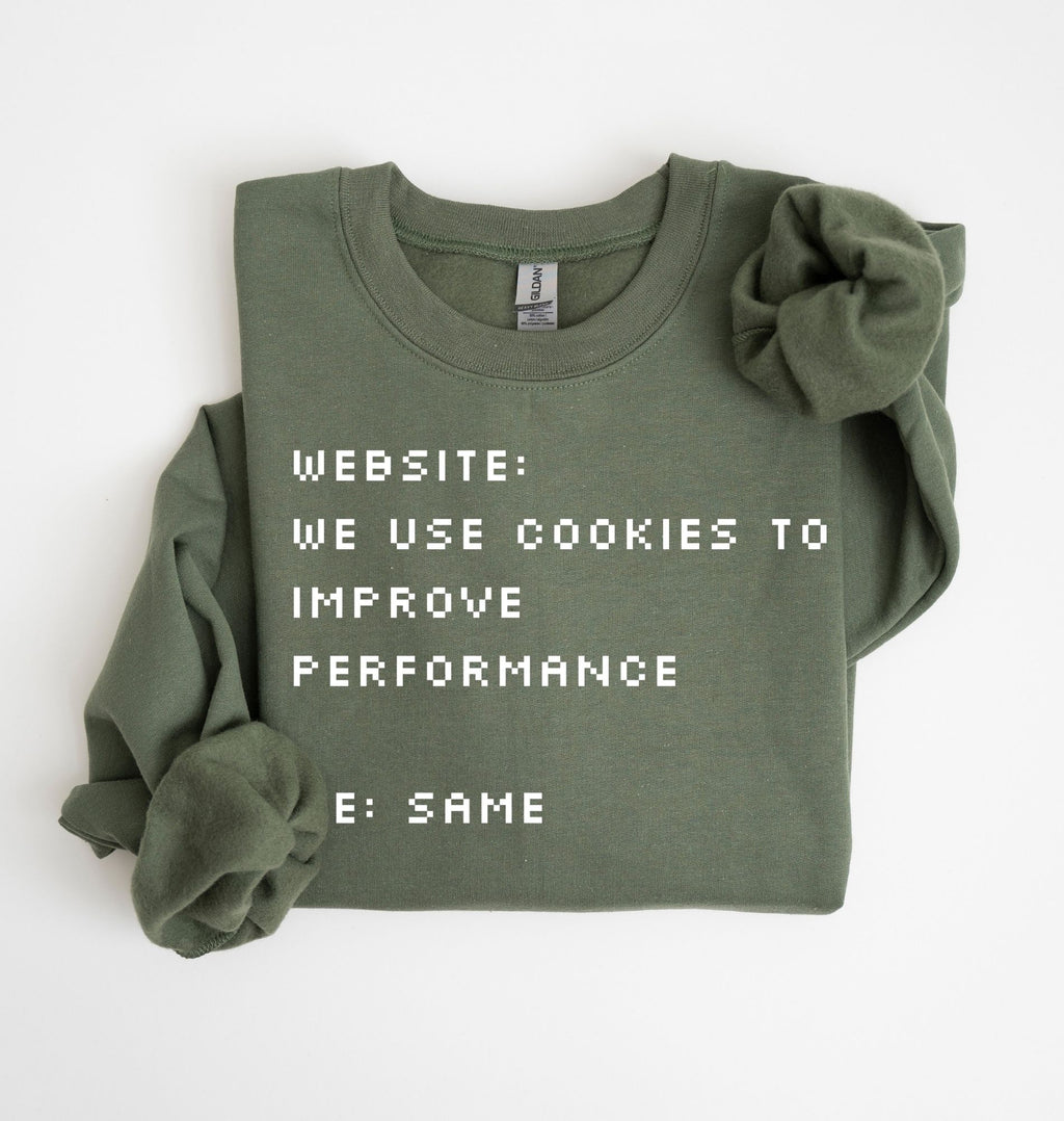 An olive green crewneck sweatshirt with a long sleeve folded over, featuring pixel-style white text that reads 'WEBSITE: WE USE COOKIES TO IMPROVE PERFORMANCE' and 'E: SAME'. A matching scrunchie is placed on the right shoulder.