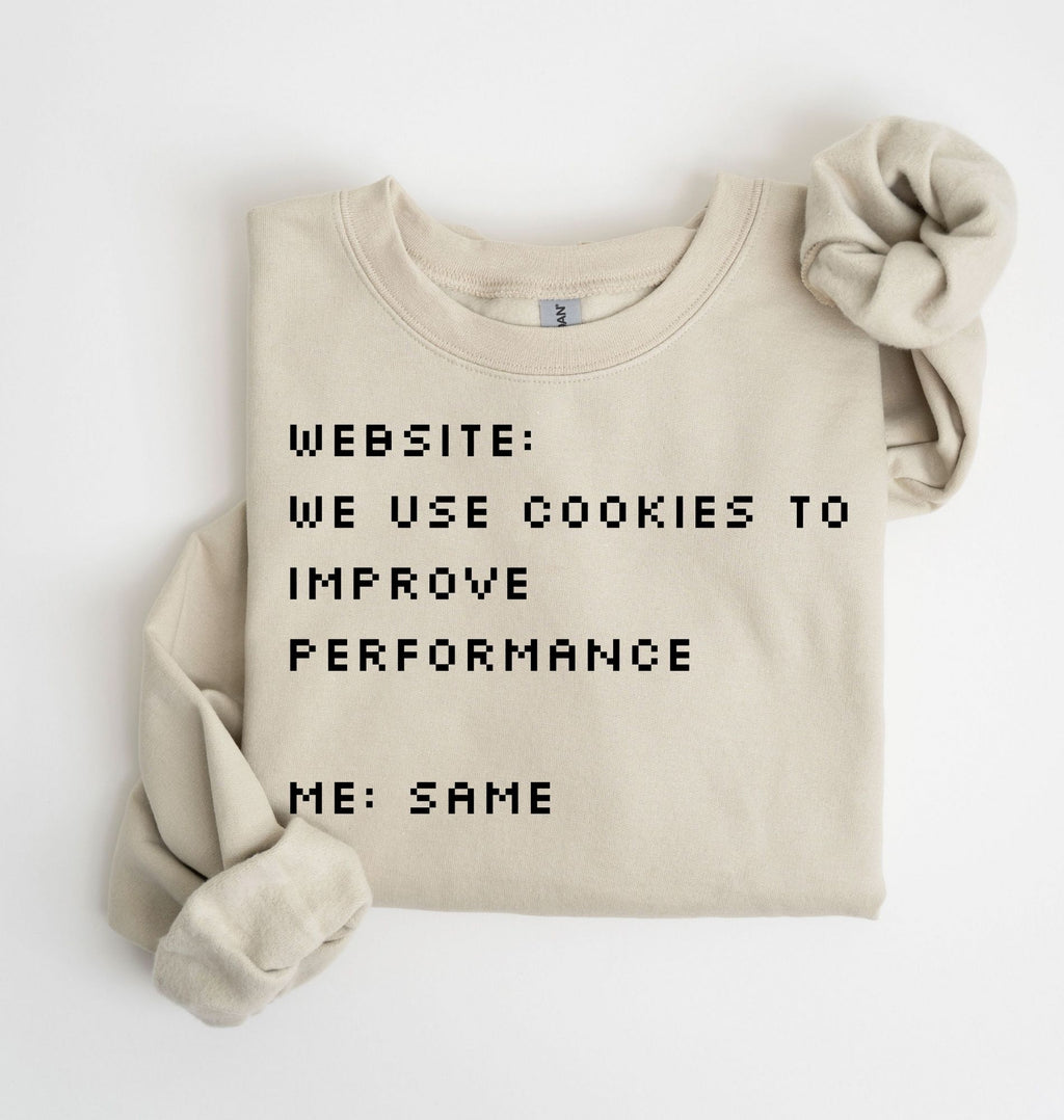A folded beige crewneck sweatshirt with black pixel-style text reading 'WEBSITE: WE USE COOKIES TO IMPROVE PERFORMANCE ME: SAME', shown on a white background with one rolled sleeve and a matching fabric scrunchie.