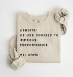 A folded beige crewneck sweatshirt with black pixel-style text reading 'WEBSITE: WE USE COOKIES TO IMPROVE PERFORMANCE ME: SAME', shown on a white background with one rolled sleeve and a matching fabric scrunchie.