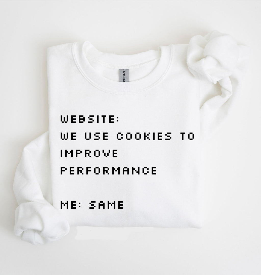 A white crewneck sweatshirt with black pixel-style text reading 'WEBSITE: WE USE COOKIES TO IMPROVE PERFORMANCE ME: SAME', shown folded on a white background with a matching white fabric prop near the shoulder.