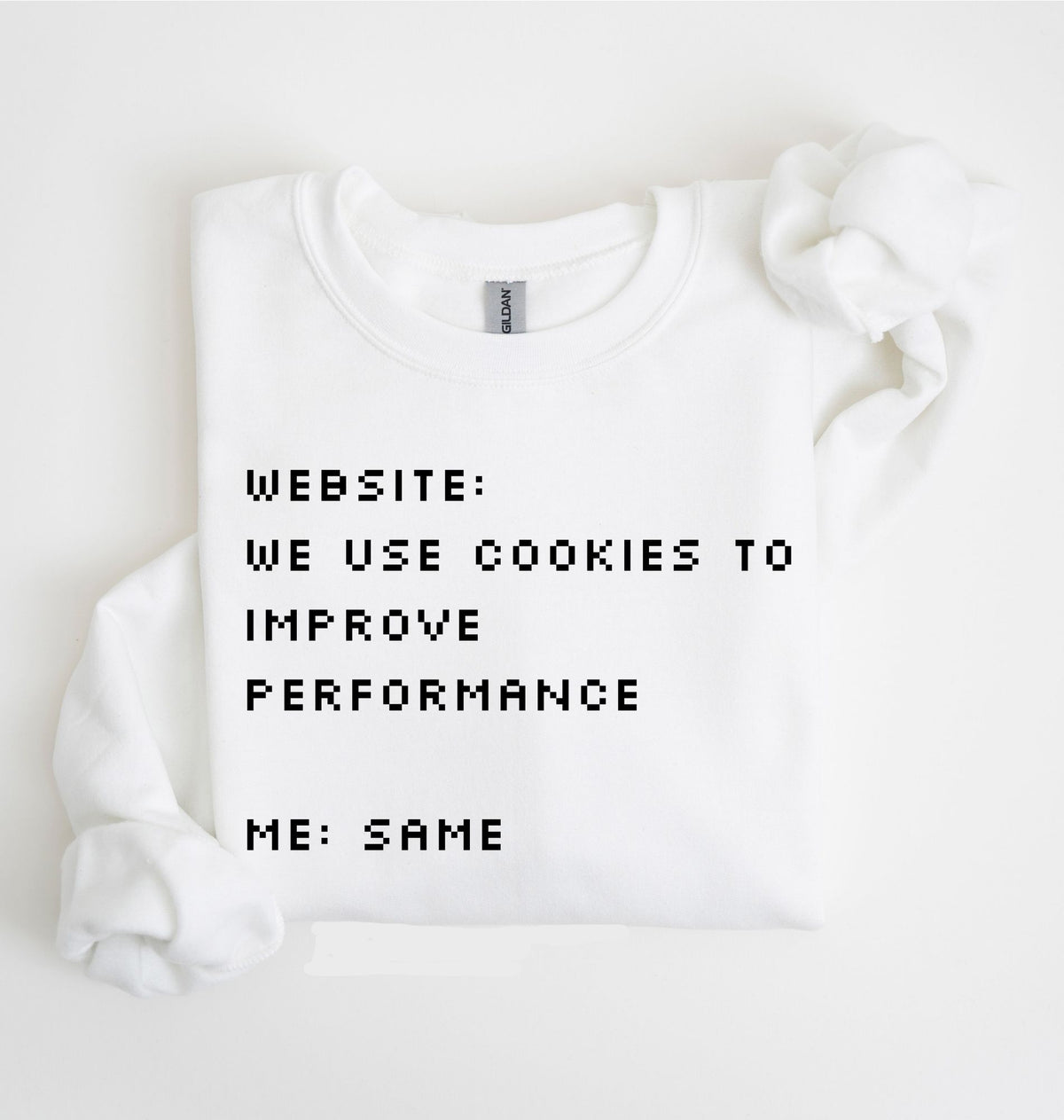 A white crewneck sweatshirt with black pixel-style text reading 'WEBSITE: WE USE COOKIES TO IMPROVE PERFORMANCE ME: SAME', shown folded on a white background with a matching white fabric prop near the shoulder.