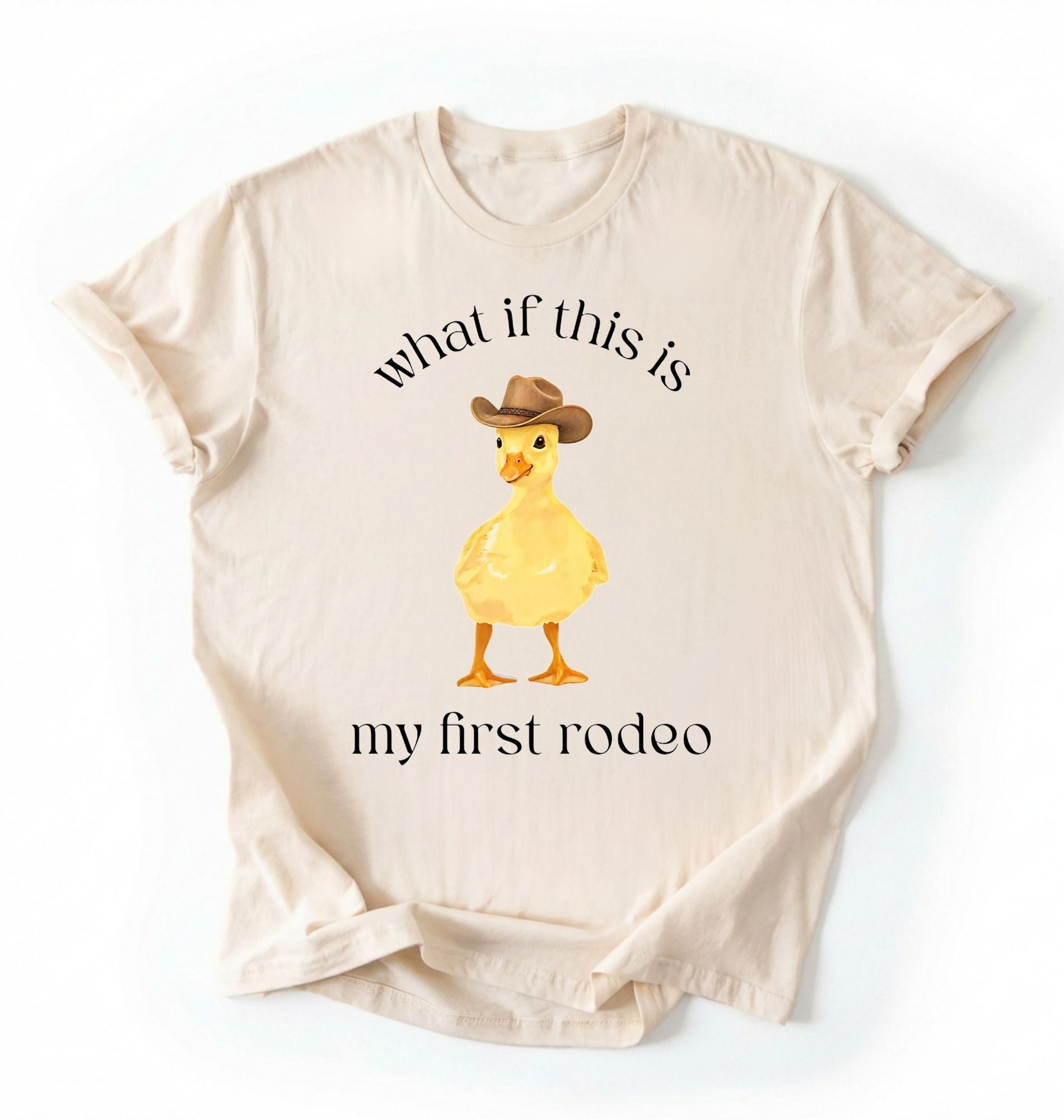 Flat lay of an ivory what if this is my first rodeo shirt brown funny cowboy duck t shirt western humor gift on a white background 