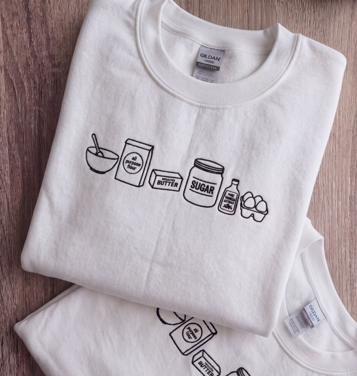 A folded white Gildan t-shirt with a black line art graphic of baking ingredients (bowl, flour, butter, sugar, syrup, eggs) on a brown wooden background.