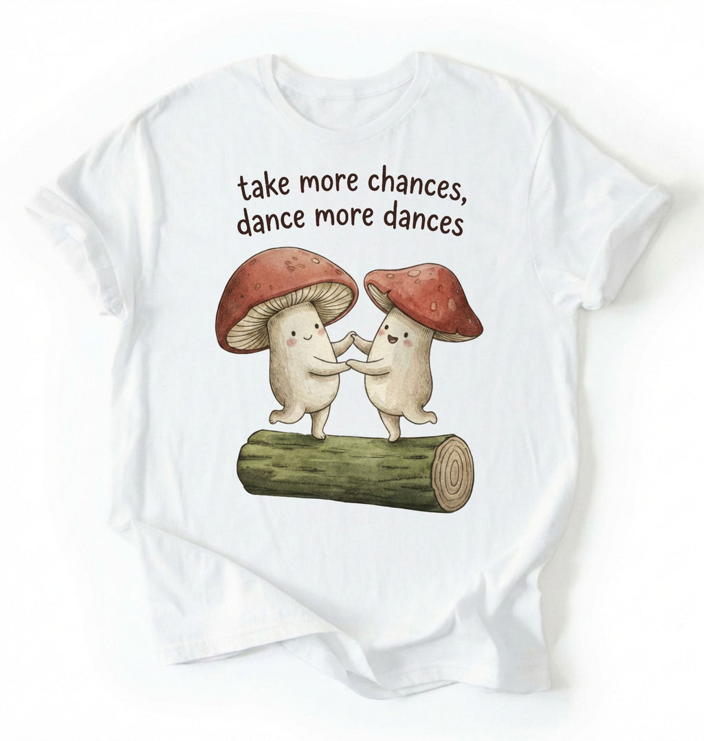 Flat lay of a White t-shirt with two dancing mushrooms and the phrase take more chances dance more dances, whimsical cottagecore gift tee on a white background 
