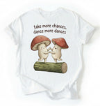 Flat lay of a White t-shirt with two dancing mushrooms and the phrase take more chances dance more dances, whimsical cottagecore gift tee on a white background 