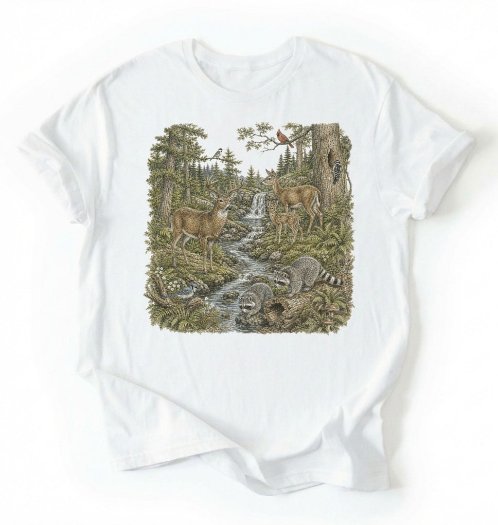 Flat lay of a white Woodland forest animals shirt featuring deer, raccoons, and scenic nature illustration on a white background 